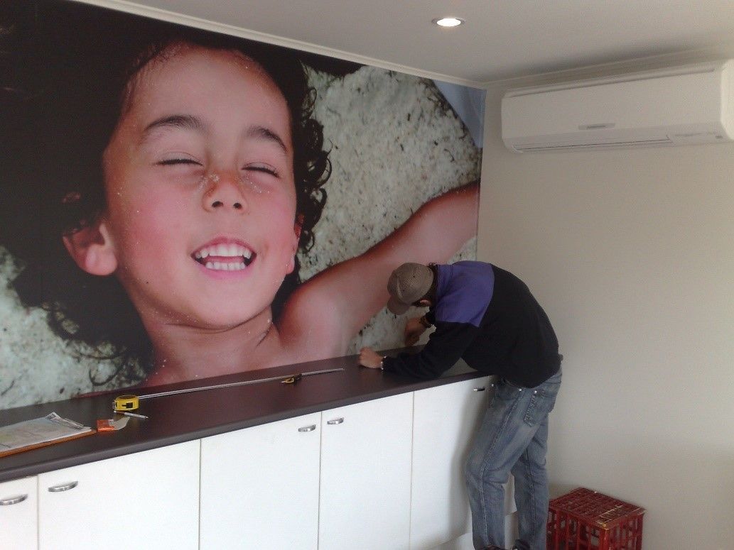 A person measures a dark countertop in front of white cabinets, with a large mural of a smiling child on the wall.